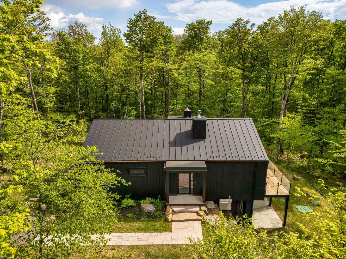 Modern black house with a metal roof nestled in a lush green forest, featuring a stone walkway and wooden deck.