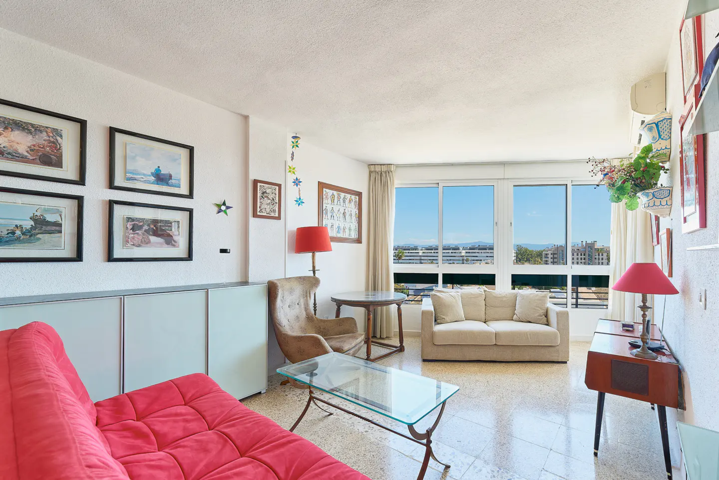 Bright living room with red sofa, beige armchair and sofa, glass coffee table, and large windows with city views.