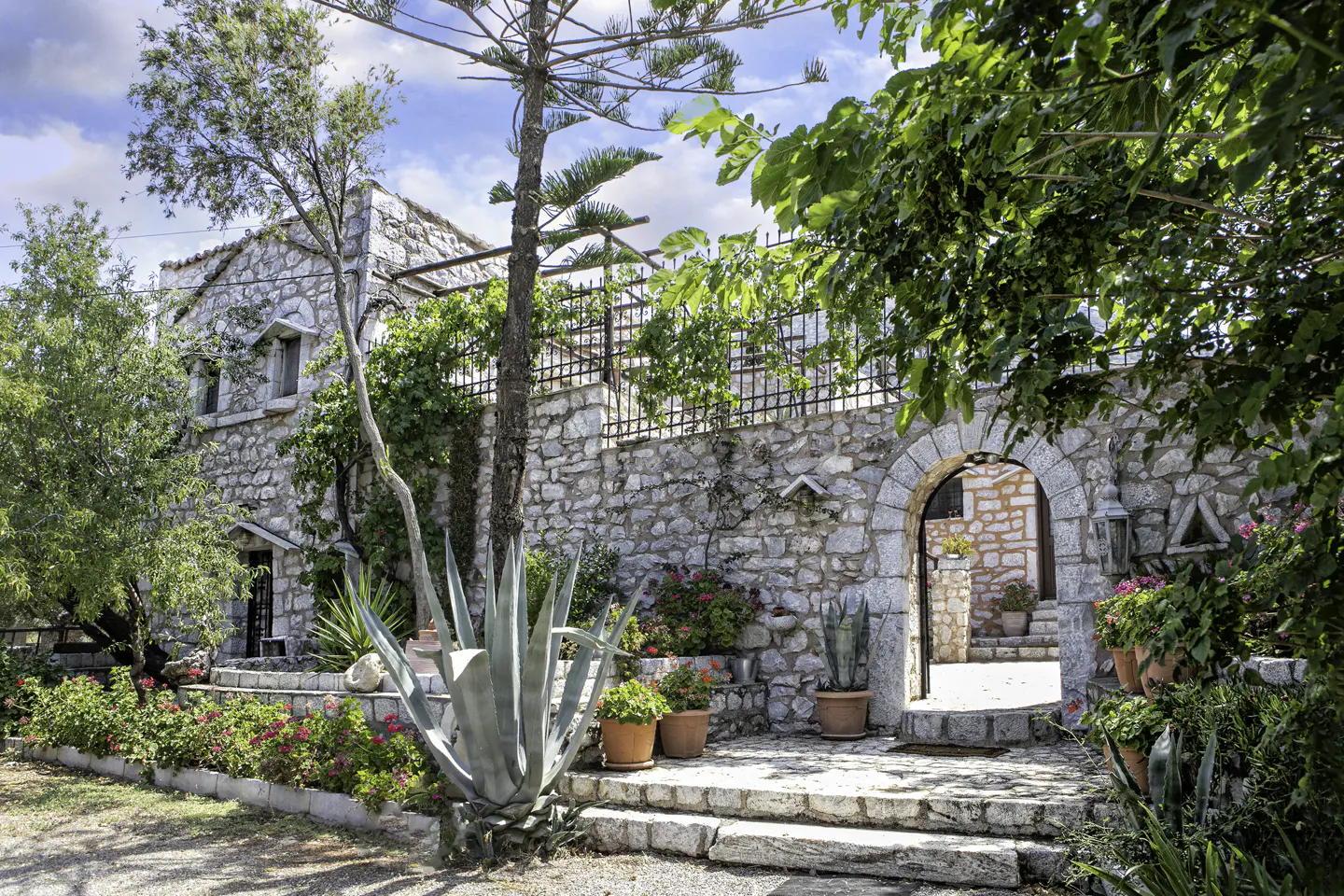 Stone house with an arched entrance and lush greenery. Flowers in pots line the steps and walls, creating a vibrant, inviting scene.