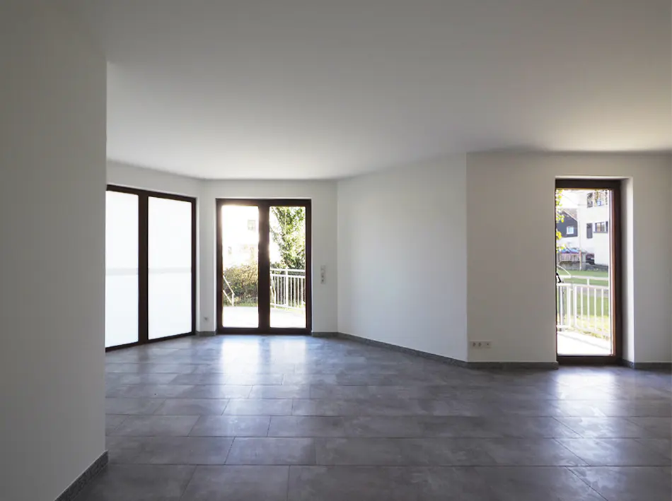 Bright, empty room with gray tile floor, white walls, and three dark-framed glass doors leading to balconies.