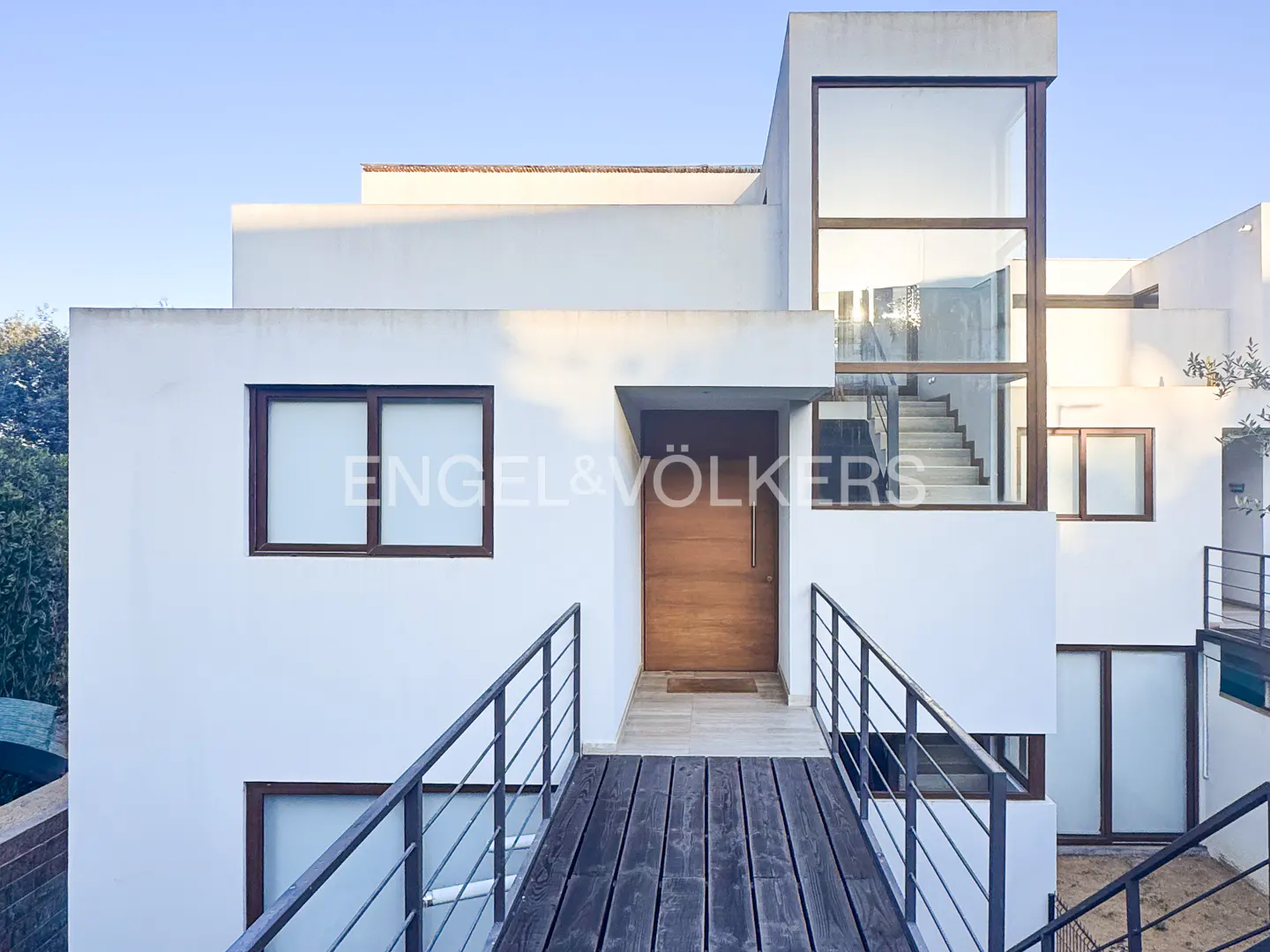 Modern white house with a wooden walkway leading to the front door. Large windows and a glass-enclosed staircase are visible.