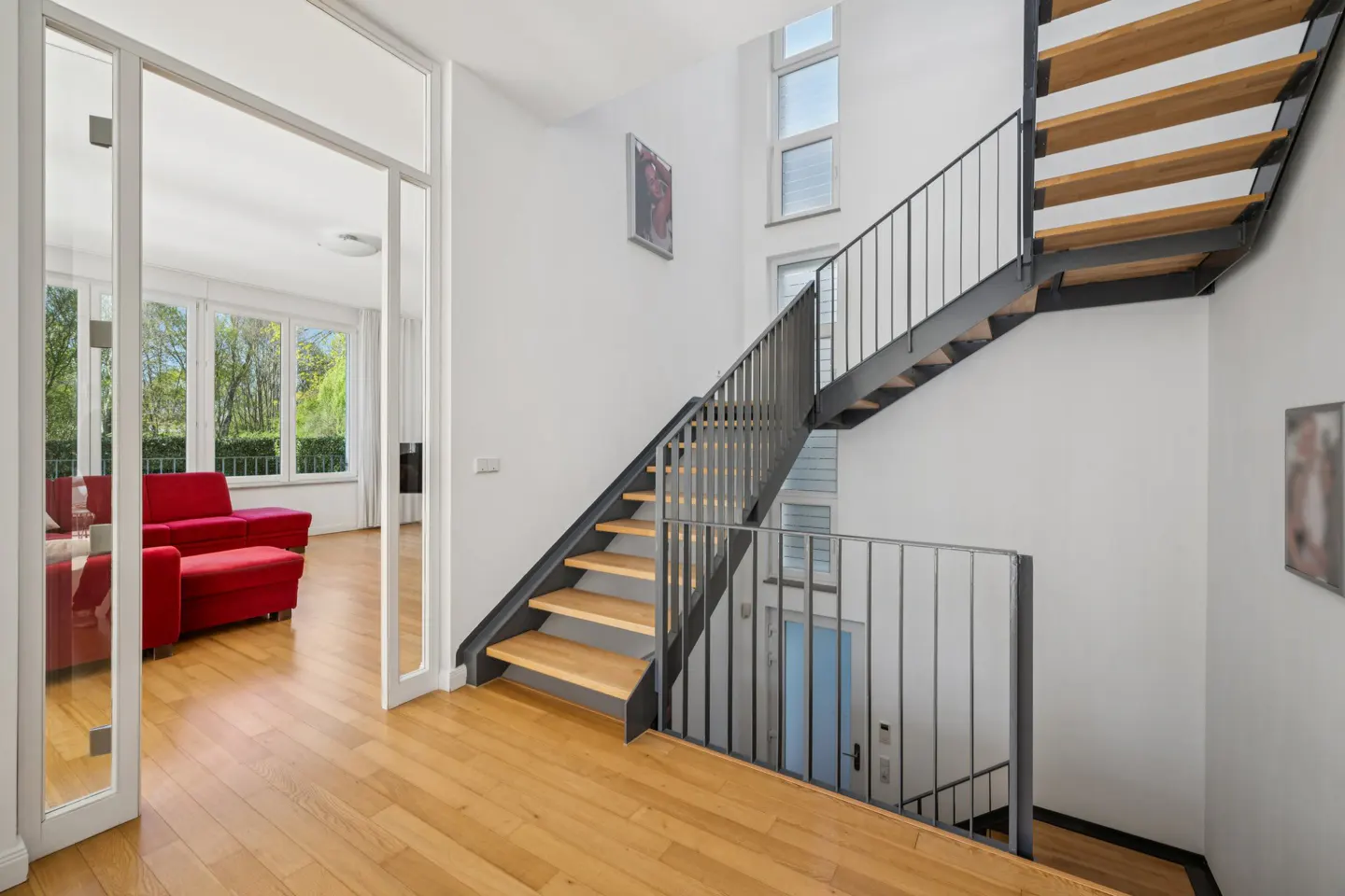 Interior view of a modern home with wooden floors, stairs with metal railings, and a red sofa visible through a glass doorway.