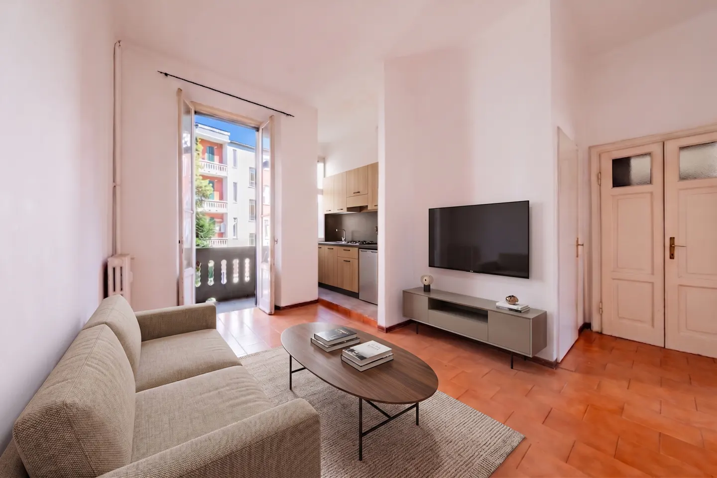 A bright living room with a beige sofa, oval coffee table, TV, and balcony with a view of a building.