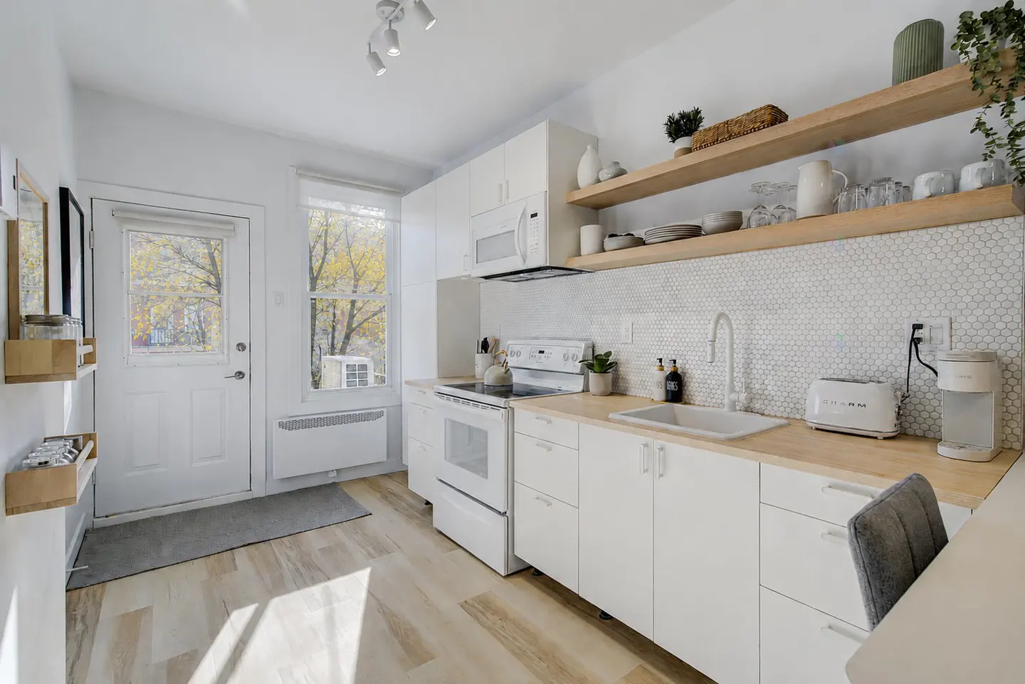 Bright, modern kitchen with white cabinets, wood countertops, and open shelves. A door leads outside, with autumn trees visible through the window.