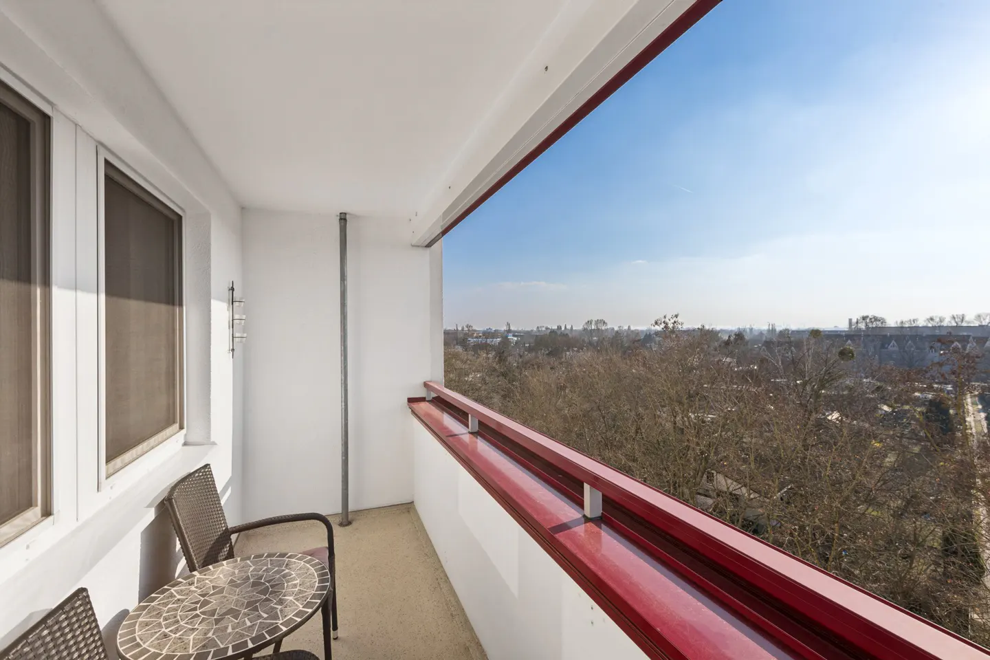 Balcony view with a mosaic table and two chairs. The red railing overlooks a cityscape under a blue sky. Windows are on the left.