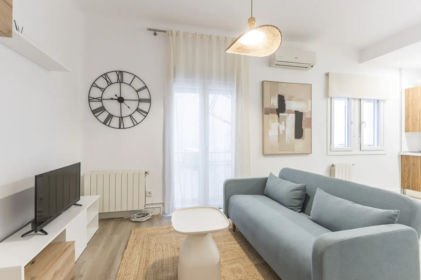 Bright living room with a light blue sofa, white walls, a large clock, and a jute rug. A white table sits in front of the sofa.