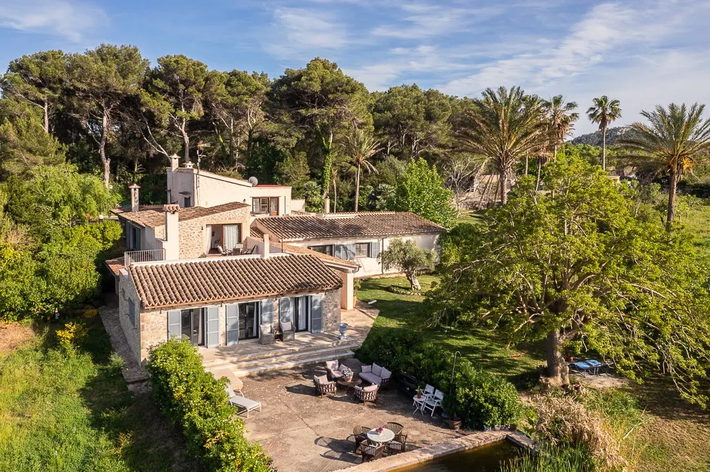 Aerial view of a stone house with a brown tile roof, patio furniture, and lush green trees.