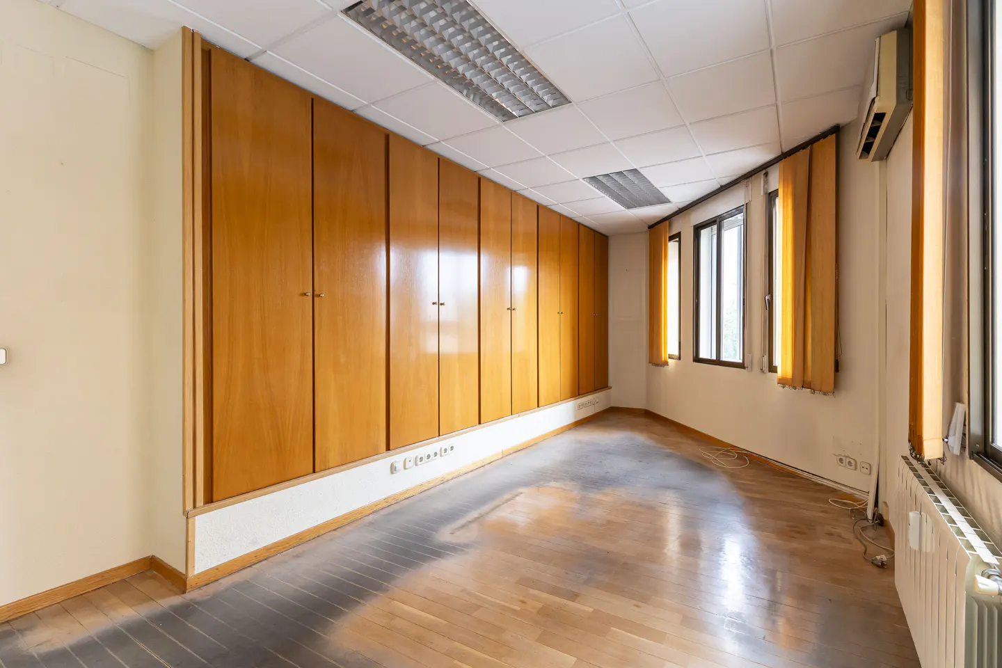 Empty room with wood floors, a wall of wood cabinets, and windows with yellow curtains.