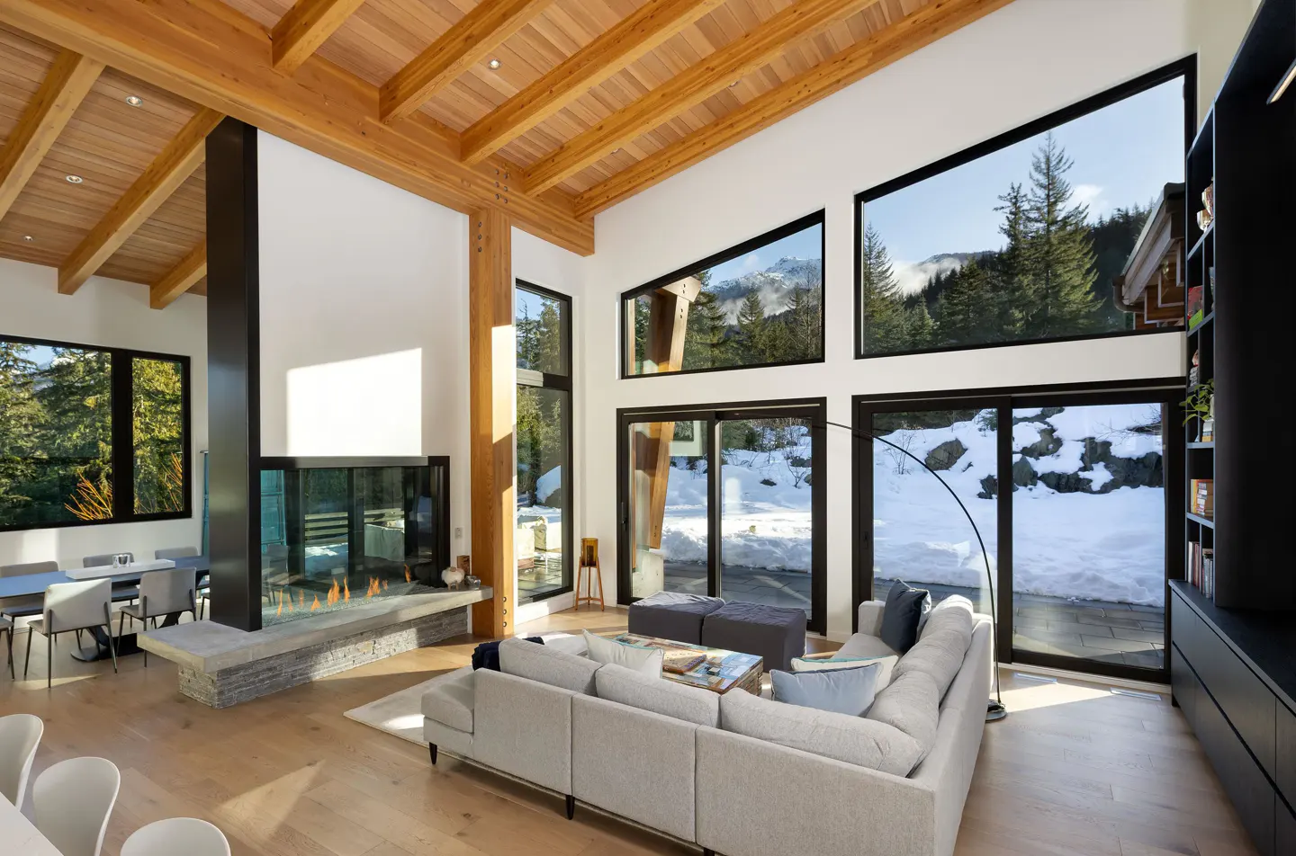 Bright living room with wood ceiling, fireplace, and large windows showing snowy mountain views. A gray sectional sofa sits on a light wood floor.