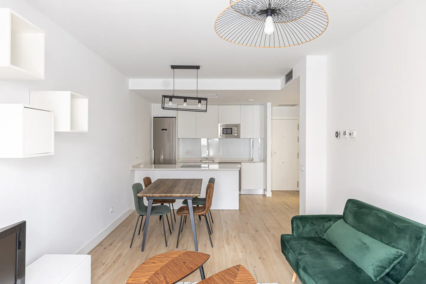 Bright, modern apartment interior with white walls, wood floors, and an open kitchen. A green sofa and wood table add color and warmth.