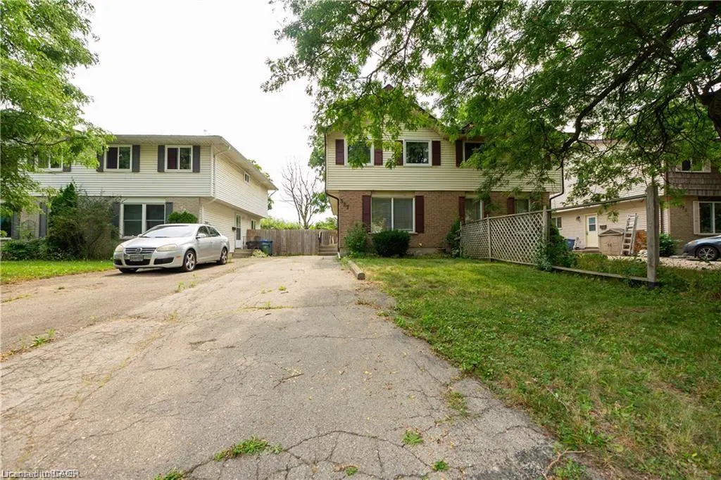 Exterior view of a two-story tan house with brick accents and maroon shutters, next to a white house with a silver car parked in the driveway.