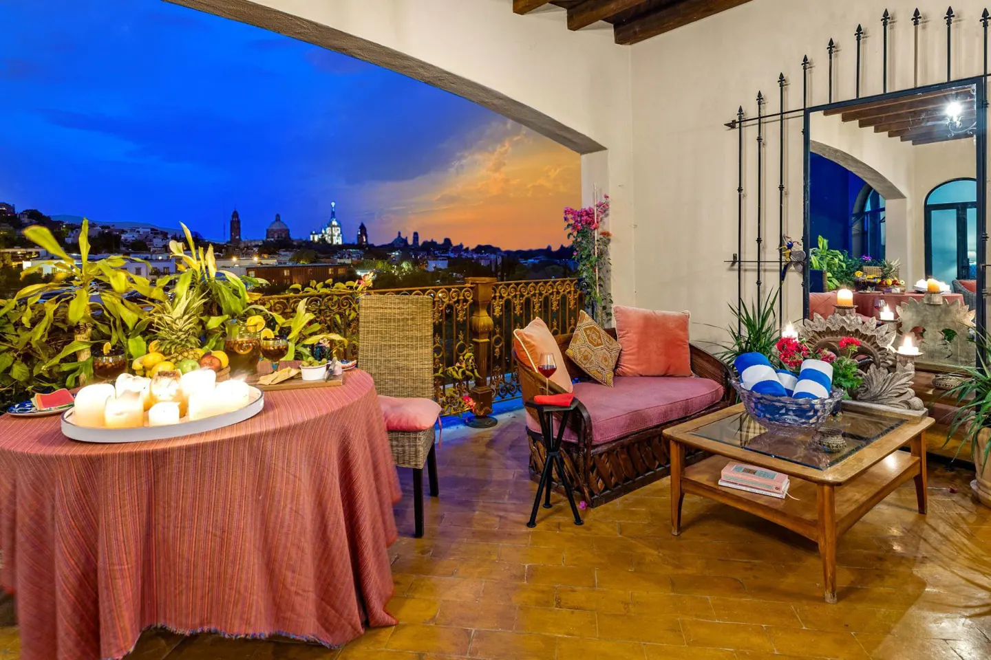 Outdoor patio with table, wicker furniture, and city view at dusk. Table has fruit, candles, and drinks. Couch has pink cushions.