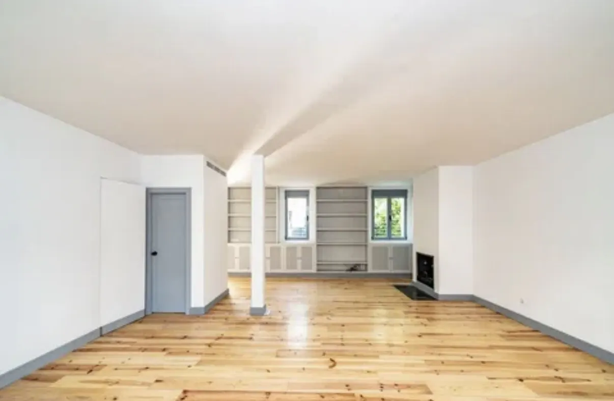 Bright, empty room with wood floors, white walls, and a gray door. Built-in bookshelves flank two windows and a fireplace.