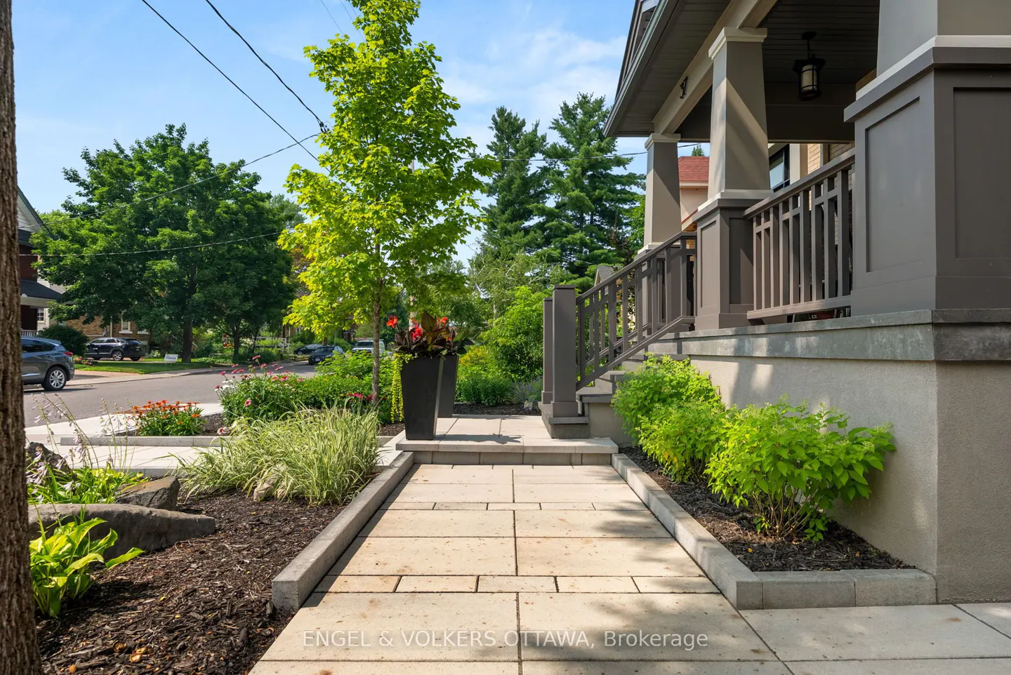 Landscaped walkway leads to a gray house with a porch and railing. Green bushes and trees surround the path.