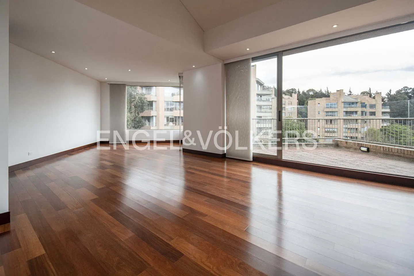 Bright, empty room with hardwood floors, white walls, and large windows overlooking a balcony and city buildings. Engel & Völkers logo visible.
