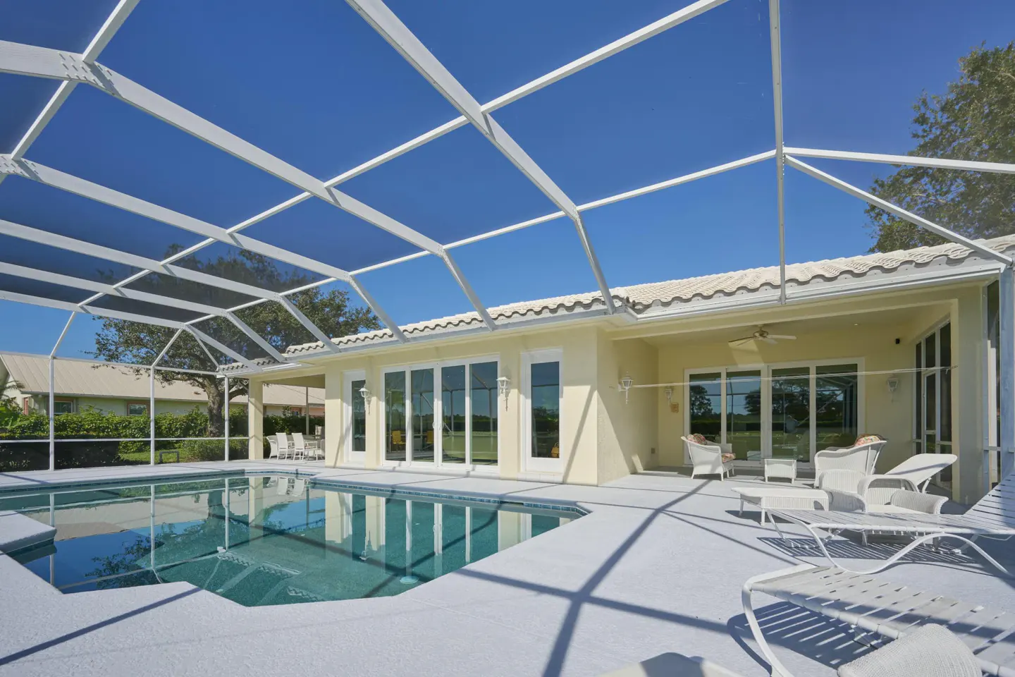 Outdoor pool area with a screened enclosure, white patio furniture, and a beige house under a clear blue sky.