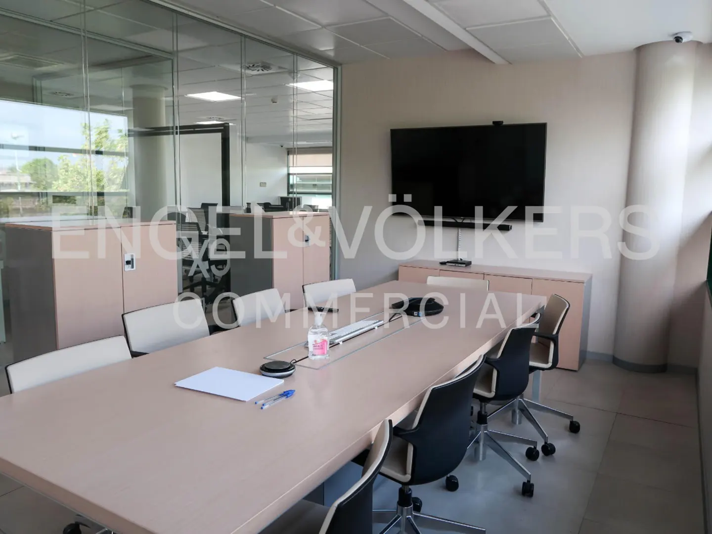 Conference room with a long table, black chairs, and a large TV screen on the wall. Glass walls and cabinets are visible.