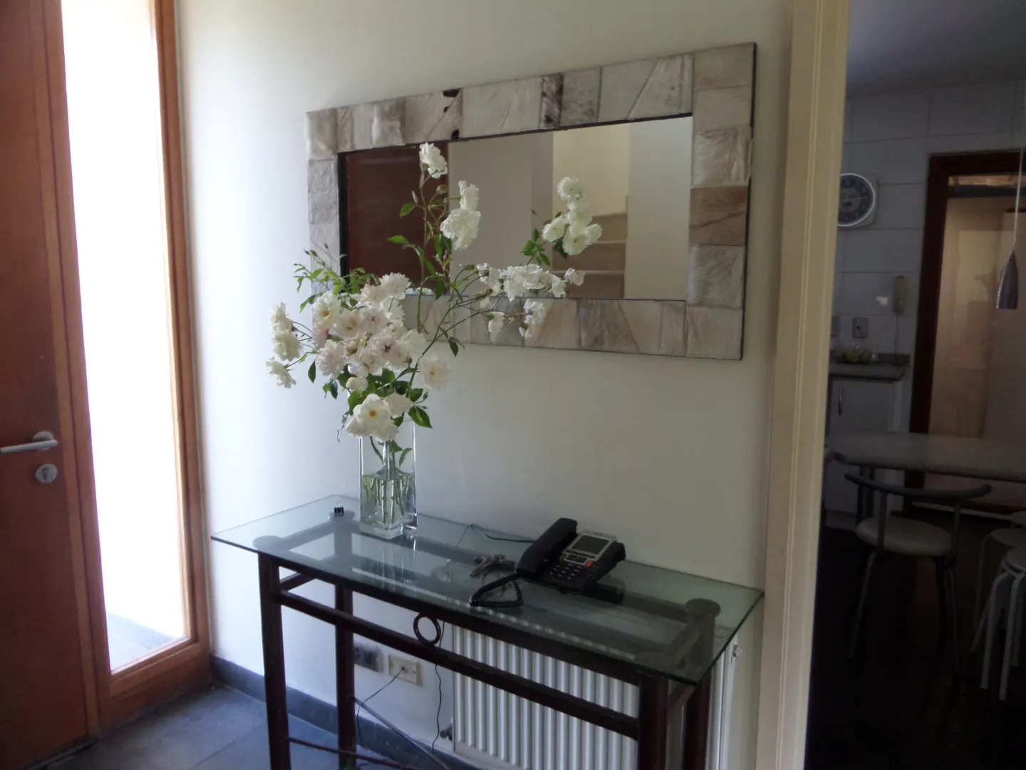 Entryway with a glass-top console table, white roses in a vase, and a black telephone. A stone-framed mirror hangs above, reflecting a staircase. A doorway reveals a kitchen.