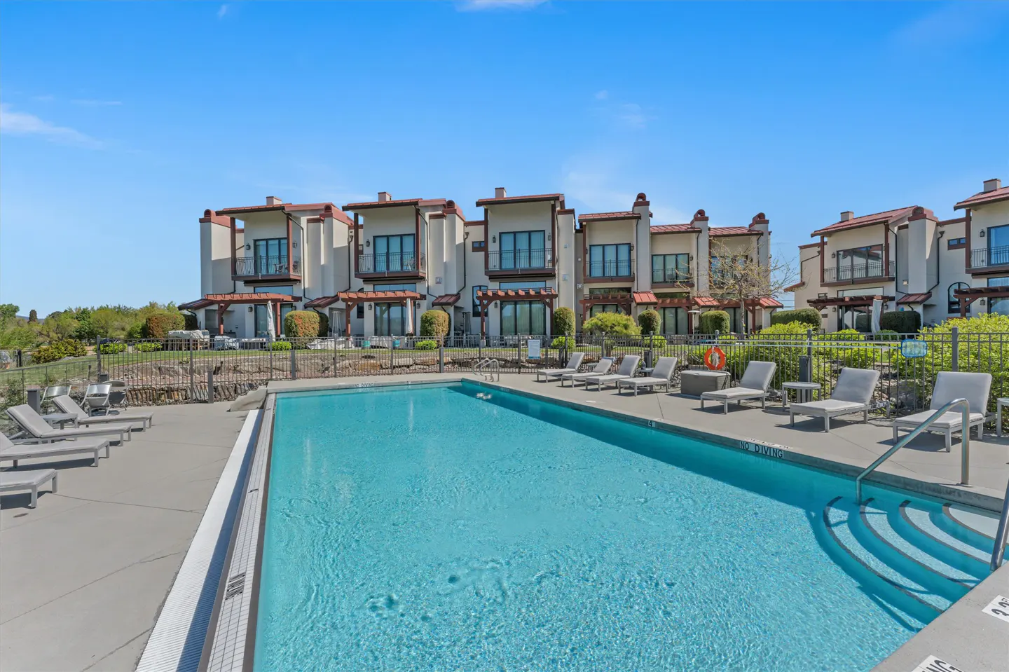 A blue swimming pool is in front of tan townhouses with lounge chairs on the pool deck.