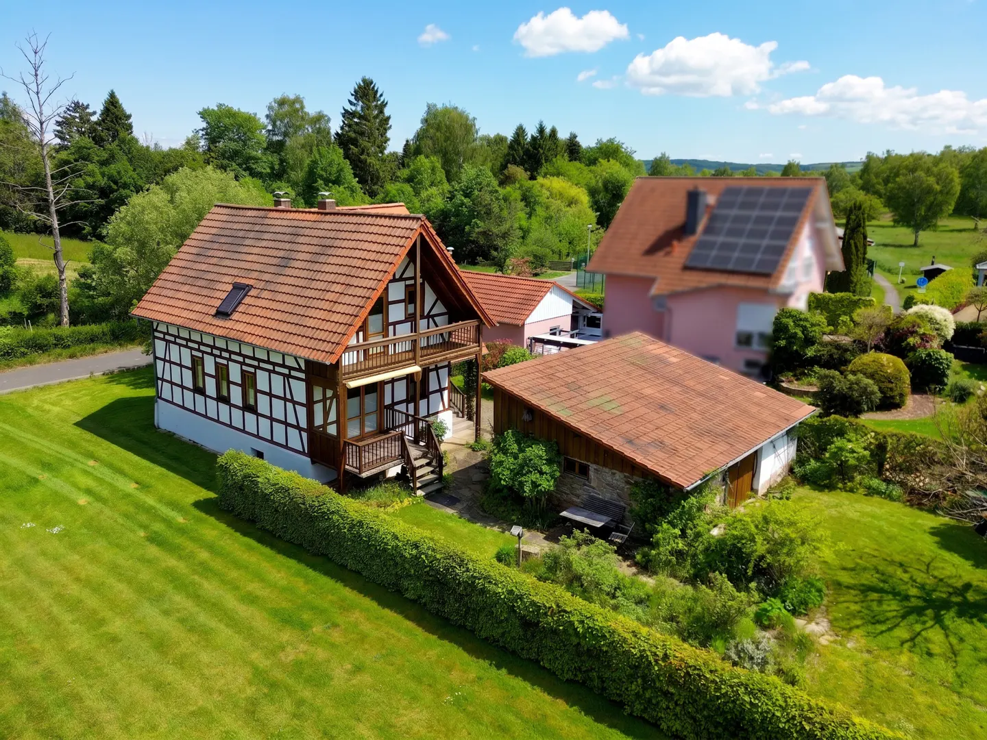 Two-story half-timbered house with a red tile roof, balcony, and green lawn. A small outbuilding and pink house are nearby.