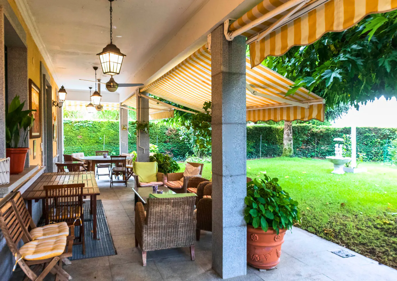 Covered patio with wicker furniture, wooden tables, and yellow striped awnings overlooking a green lawn with a fountain.