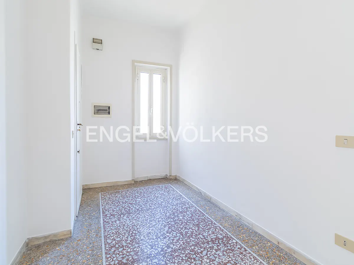 Bright, empty room with white walls, a window, and a patterned tile floor. An Engel & Völkers logo is visible on the wall.