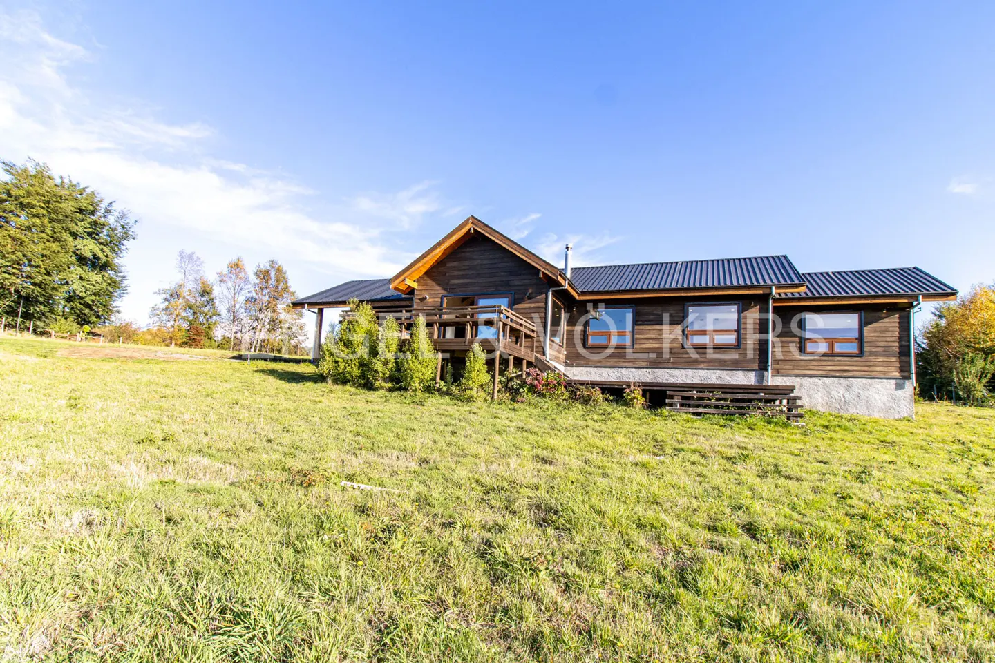 A rustic brown wooden cabin with a dark metal roof sits on a grassy hillside under a clear blue sky.