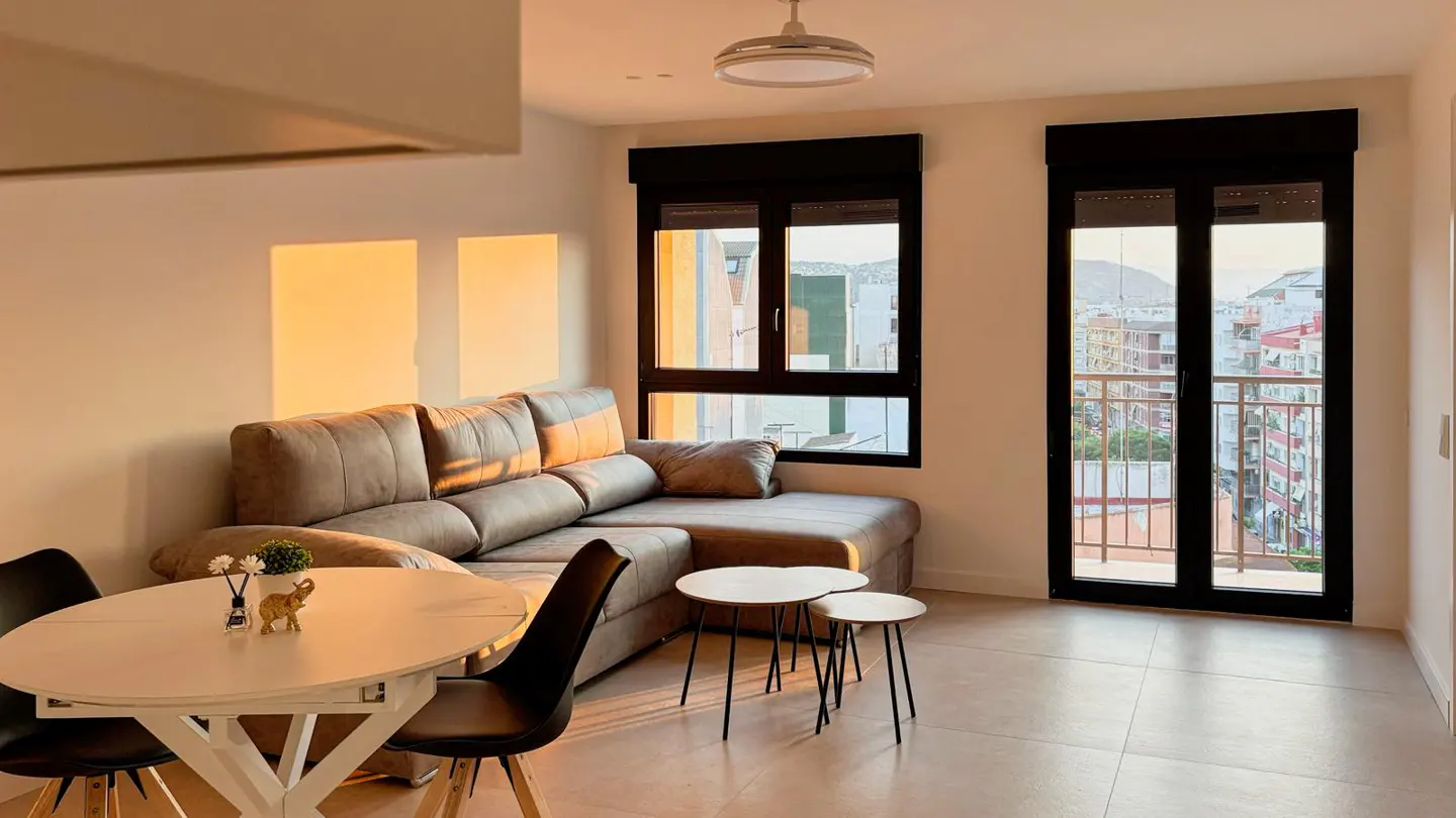 Bright apartment interior with a gray sectional sofa, white table and chairs, and a view of the city through black-framed windows.