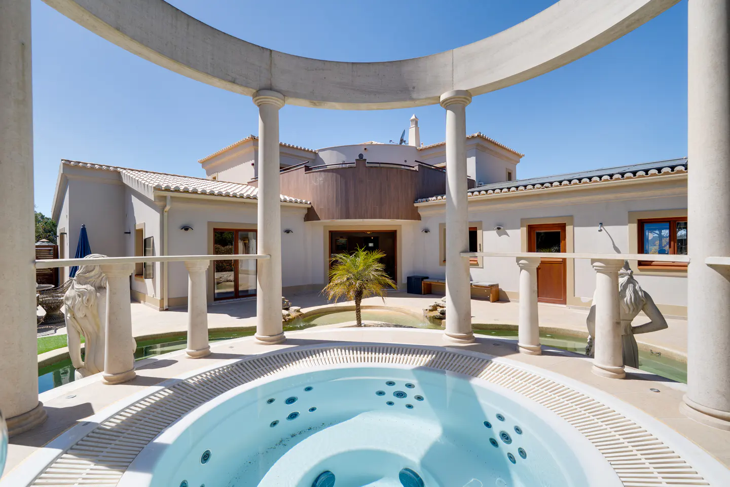 Outdoor jacuzzi with white columns and statues, set against a large beige house with a palm tree in the courtyard.