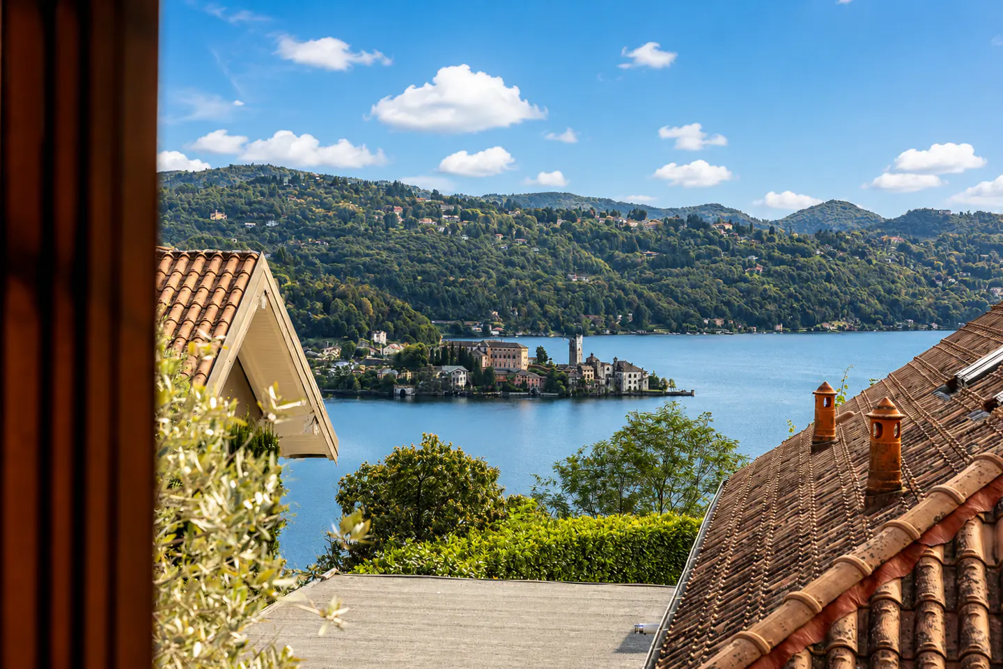 View of Lake Orta, Italy, from a window. Red tile roofs frame the blue water and island with buildings. Green hills and a blue sky with clouds are in the background.