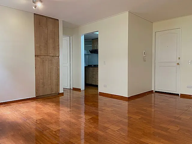 A bright, empty living room with shiny wood floors, white walls, and a wood cabinet. A doorway leads to the kitchen. A white door is on the right.