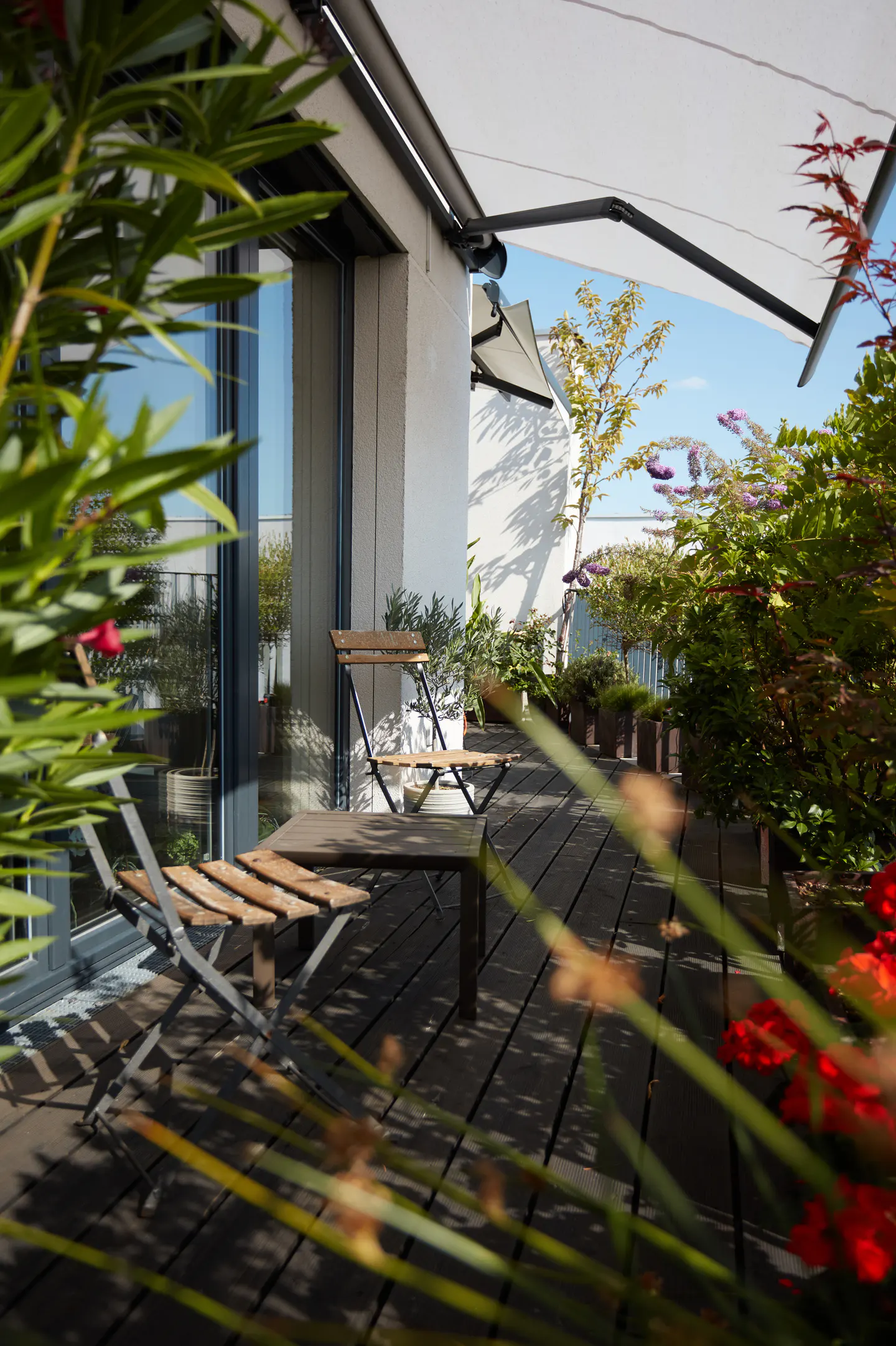 A rooftop patio with a wooden deck, table, chairs, and potted plants under a white awning.