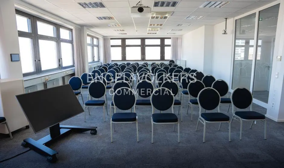 Bright conference room with rows of blue chairs, a projector, and a large screen on wheels.