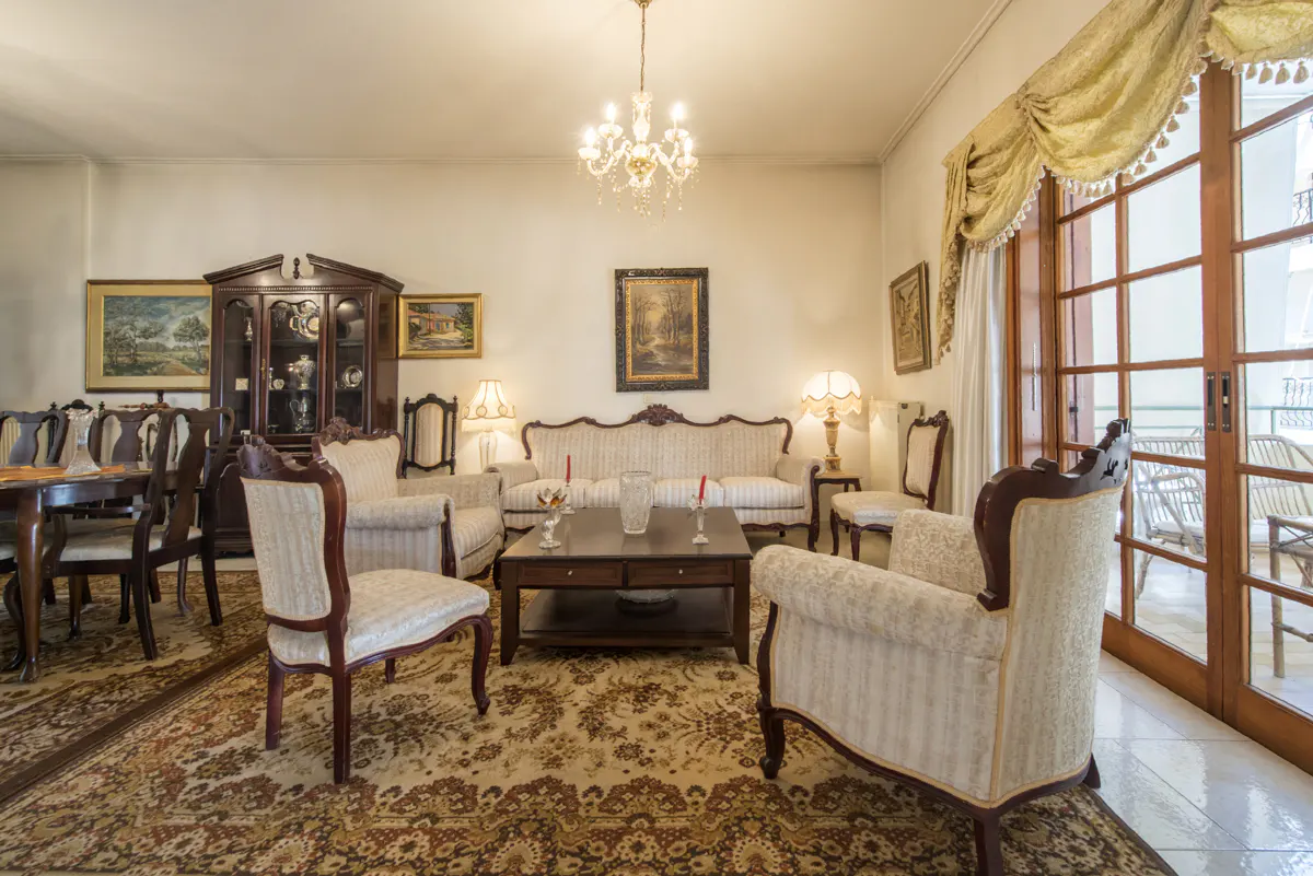 Living room with antique furniture, including a china cabinet, sofa, chairs, and a chandelier. A patterned rug covers the floor.