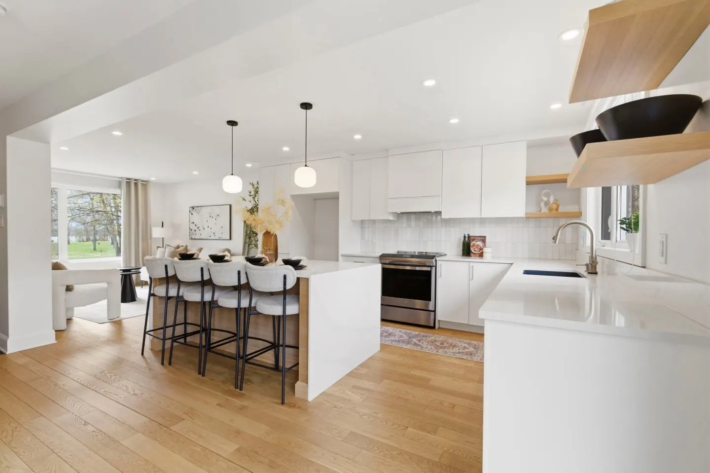 Bright, open-concept kitchen with white cabinets, island with bar stools, and light wood floors. Living room visible in the background.