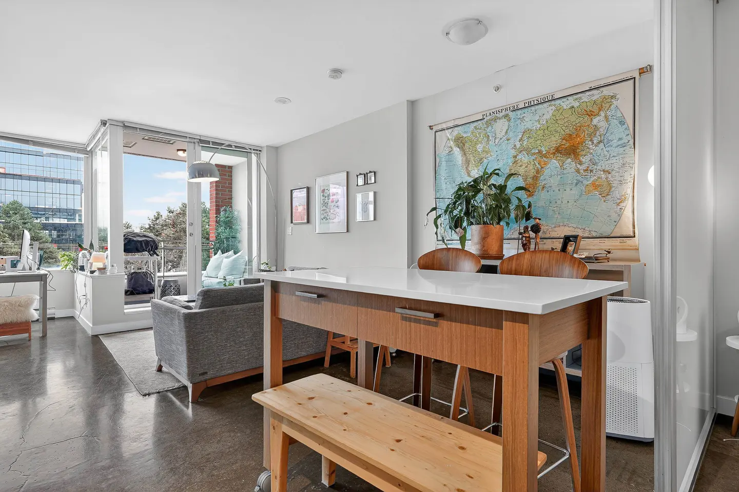 Bright, modern apartment interior with a gray sofa, wood table, and a large world map on the wall. Balcony with city views visible.