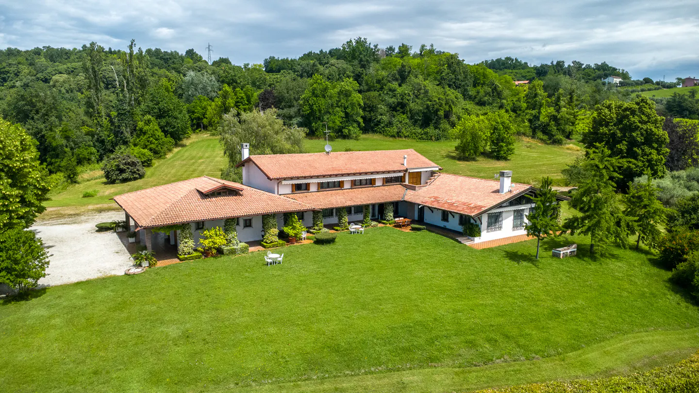 Aerial view of a white villa with a terracotta roof, surrounded by a large green lawn and lush forest under a cloudy sky.