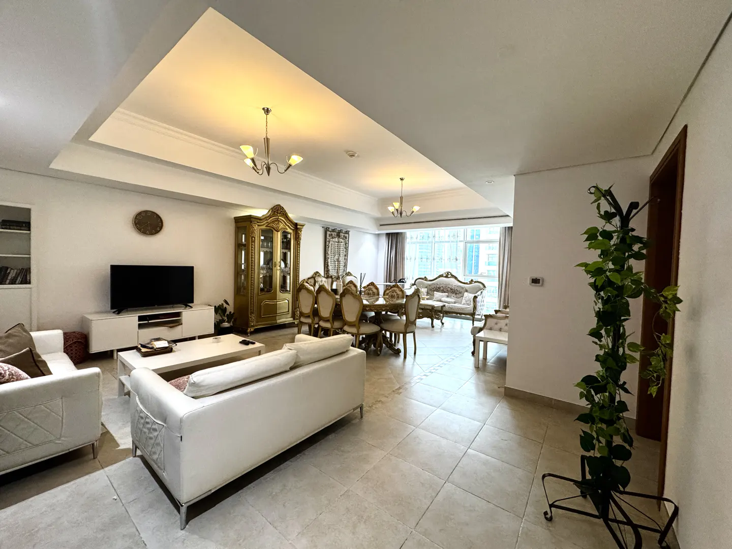 A wide shot of a bright, open-concept living and dining area with white sofas, a dining table, and a gold display cabinet.