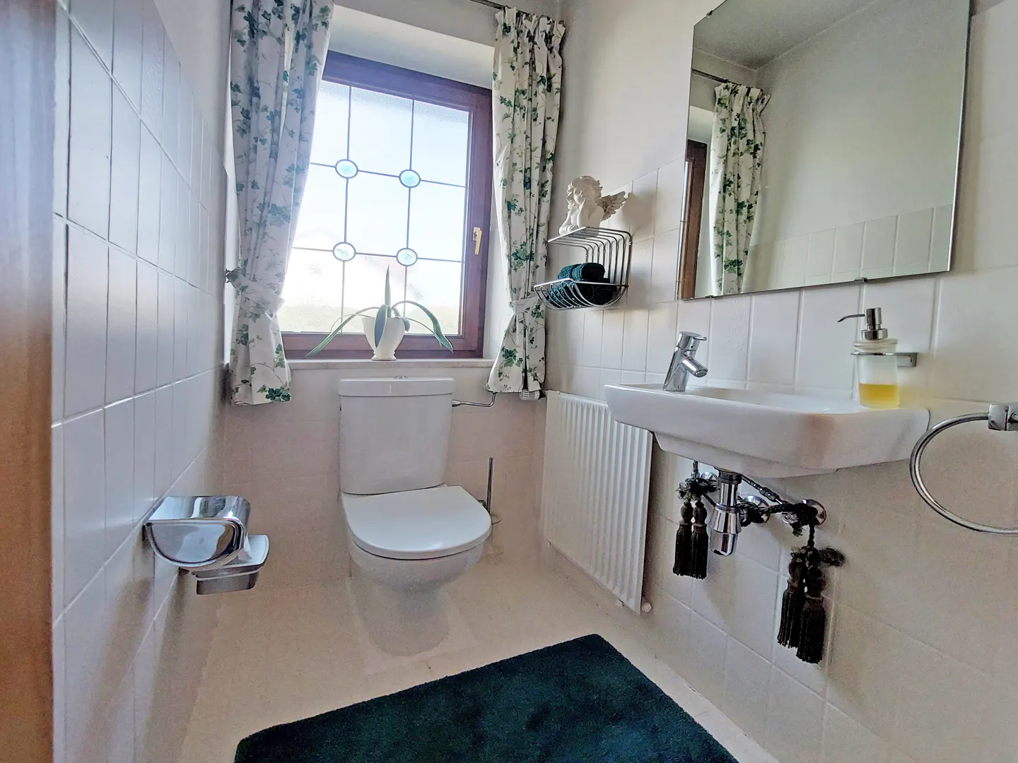 A small bathroom with white tile walls, a toilet, a sink, and a window with floral curtains.