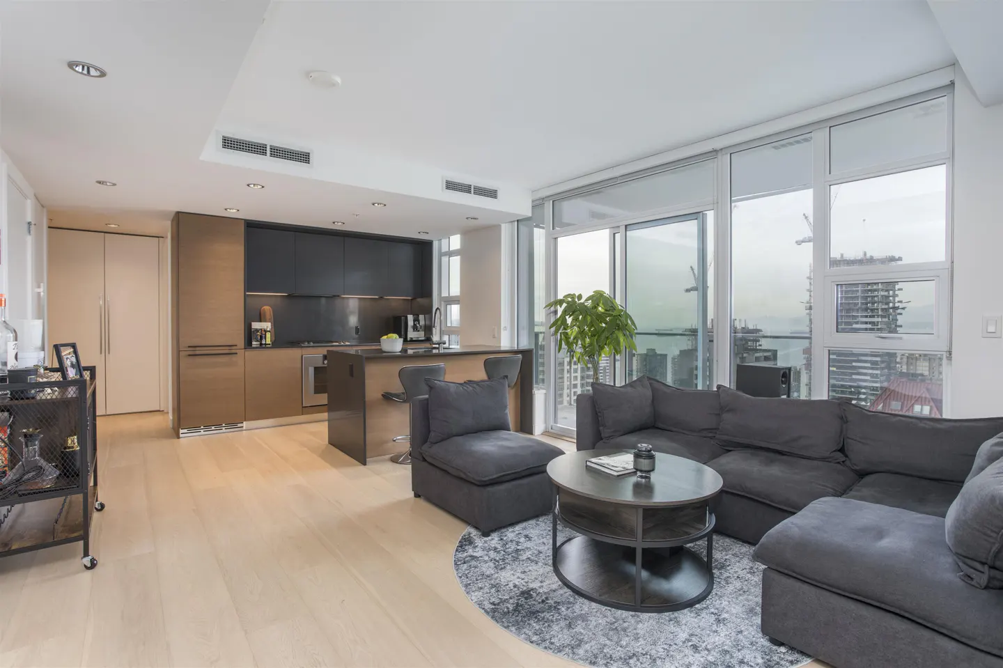 Open-concept condo with wood floors, a gray sectional sofa, and a kitchen with dark cabinets. Large windows offer city views.