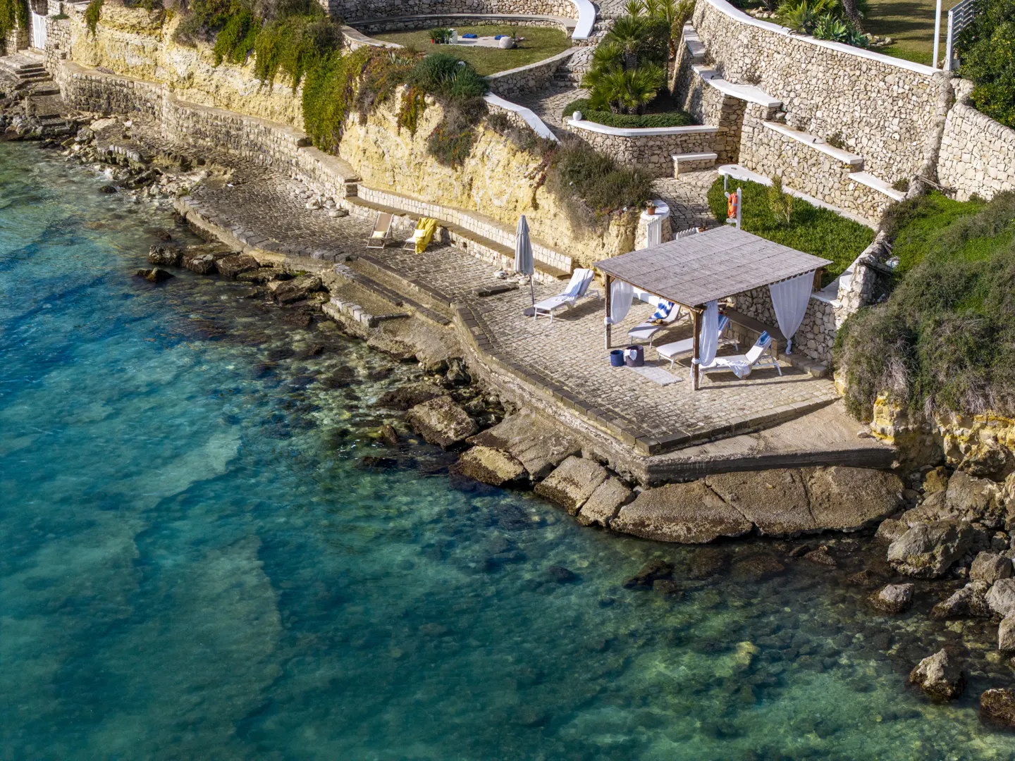 Aerial view of a rocky coastline with turquoise water, a stone patio with a gazebo, and lounge chairs.