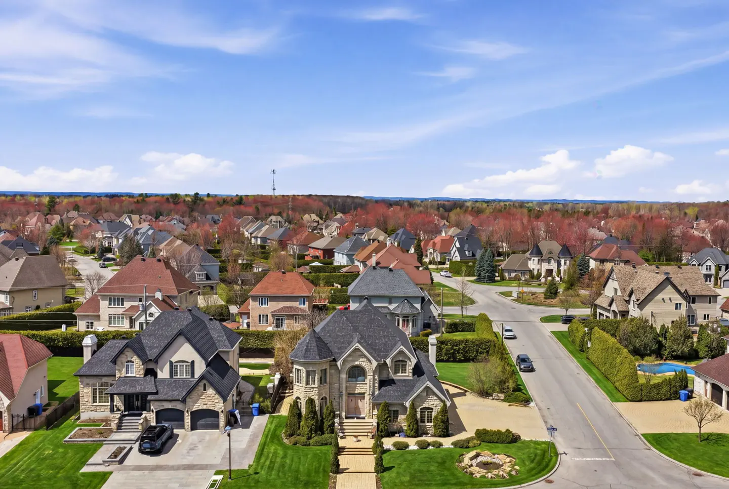 Aerial view of a suburban neighborhood with large houses, green lawns, and a blue sky with scattered clouds.