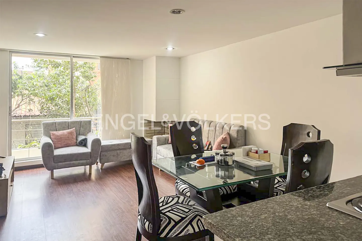Living room with gray sofa, chair, and glass table with black chairs. Large window with trees outside.