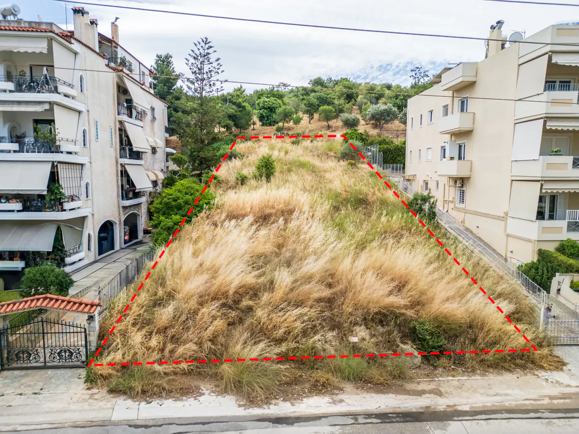 A vacant, grassy plot of land outlined in red, situated between two multi-story residential apartment buildings.