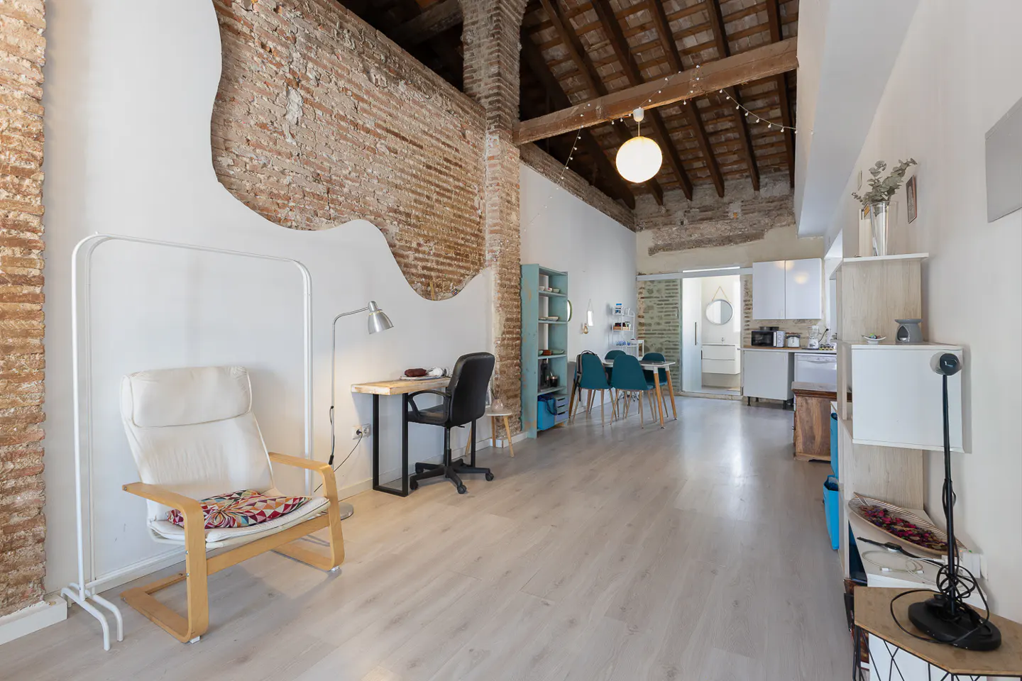Bright, open apartment with exposed brick, white walls, and light wood floors. A desk, chair, and dining table are visible.