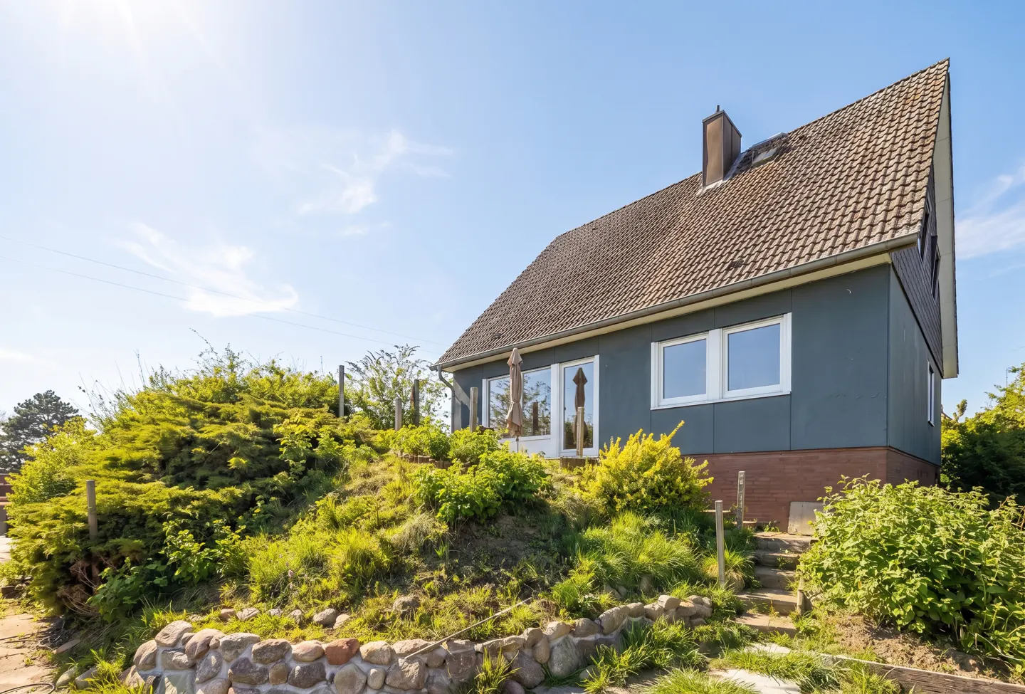 Exterior view of a blue house with a brown roof, set on a grassy hill under a clear blue sky.
