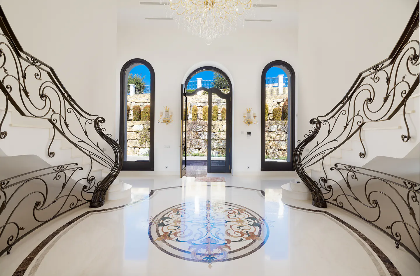 Grand foyer with marble floors, ornate staircase, and arched doorway leading to a stone garden. A crystal chandelier hangs above.
