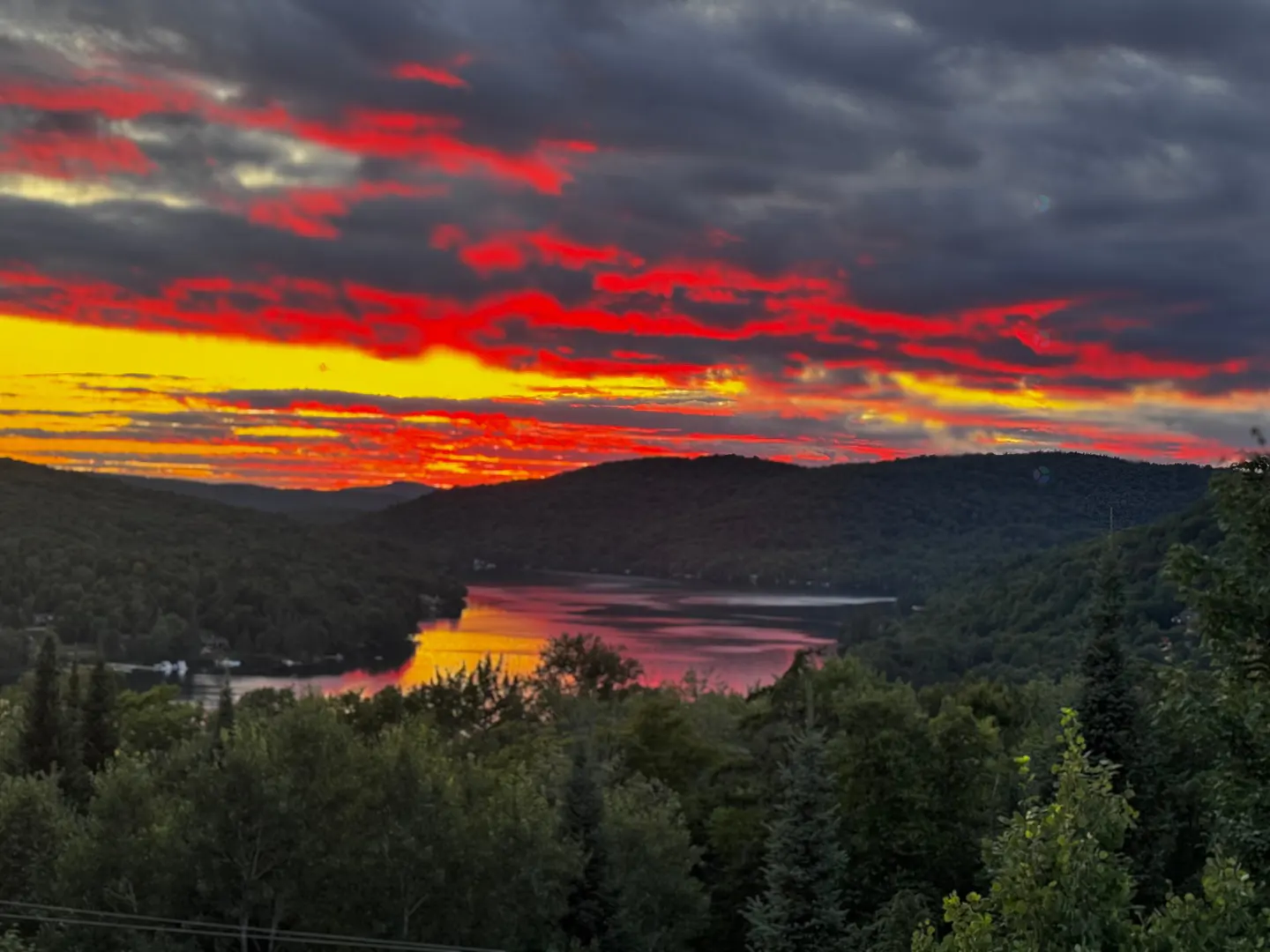 Sunset view of a lake surrounded by green hills. The sky is filled with red and yellow clouds.