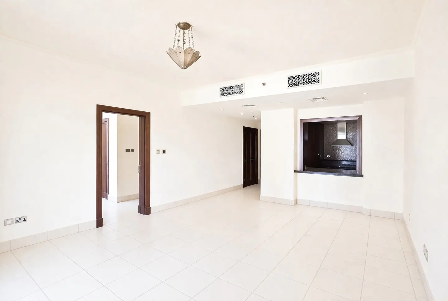 Bright, empty room with white walls and tiled floor. A doorway and kitchen pass-through are visible. A chandelier hangs from the ceiling.
