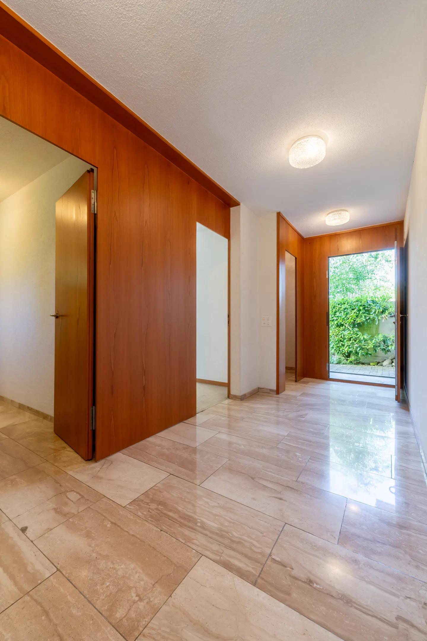 A bright, empty room with marble floors, wood paneling, and an open door to a green garden.